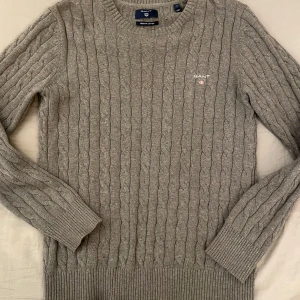 Grå kabelstickad tröja från GANT - Säljer en stilren grå kabelstickad tröja från GANT i premium bomull. Tröjan har en klassisk rund hals och broderad logga på bröstet. Perfekt för kyligare dagar! Modellen är 177 cm lång och väger 70 kg