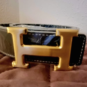 Svart bälte med guldfärgat spänne - Snyggt svart bälte med ett stort guldfärgat H-spänne. Perfekt för att ge en lyxig touch till din outfit. Bältet har vita sömmar som ger en fin kontrast.