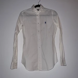 Vit skjorta från Polo Ralph Lauren - Krispig vit skjorta från Polo Ralph Lauren i slim fit. Skjortan har långa ärmar och är dekorerad med det klassiska blå broderade logotypen på bröstet. Perfekt för en stilren look. Helt kritvit, tog bild med blixt därför kan färgen se lite grå ut. Aldrig använd. 