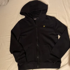 Svart Lyle&Scoot hoodie med dragkedja - Snygg svart hoodie från Lyle & Scott med dragkedja och en liten gul logga på bröstet. Perfekt för en avslappnad stil. Den har långa ärmar och en praktisk huva.