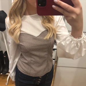 Vit och rutig blus med puffärmar - Snygg blus med vit överdel och rutigt mönster i grått och beige. Blusen har långa puffärmar med elastiska ärmslut och en figurnära passform. Perfekt för en stilren look.