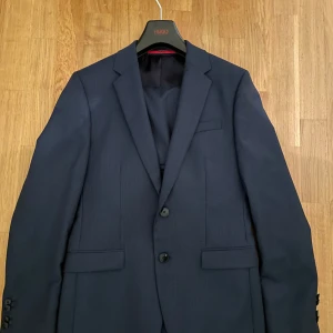 Mörkblå kostym från Hugo Boss - Snygg mörkblå kavaj från Hugo med två knappar framtill och klassiska slag. Kavajen har en elegant insida med röda detaljer och flera innerfickor. Storlek 44 (passar XS-S)