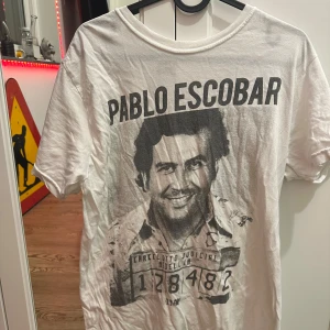Vit t-shirt med tryck - Vit t-shirt med ett stort tryck av en person och texten 'Pablo Escobar' på framsidan. T-shirten har korta ärmar och en rund halsringning. Perfekt för en avslappnad stil.