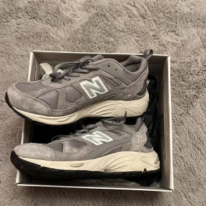 Grå sneakers från New Balance - Snygga grå sneakers från New Balance med vit sula och klassisk N-logga på sidan. Skorna har snörning och är tillverkade i en kombination av mocka och syntetmaterial för en stilren look. Jag kan stå för frakten osv! 