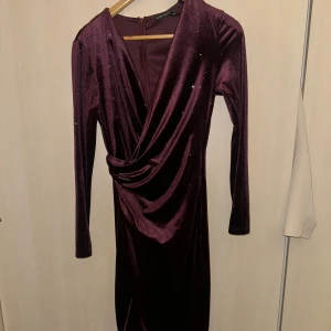 Vinröd klänning med omlott och glittriga detaljer - Elegant vinröd klänning med omlott-design och glittriga detaljer. Klänningen har långa ärmar och är tillverkad i ett mjukt material som ger en lyxig känsla.