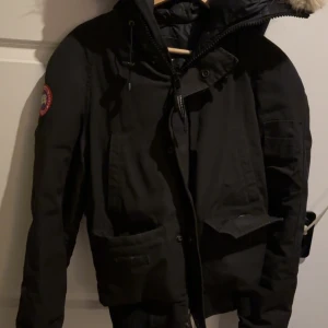 Äkta svart canada goose jacka - Säljer en svart vinterjacka från Canada Goose med pälsdetaljer på huvan. Jackan har en dragkedja och knappar framtill samt en ikonisk logga på ärmen. Perfekt för kalla vinterdagar. Den är Xs men passar som S 
