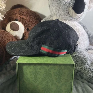 Svart keps från Gucci - Går att förhandla/pruta! Snygg svart keps från Gucci med det ikoniska mönstret och en röd-grön randdetalj på sidan. Kepsen har justerbar passform med ett bälte baktill och är perfekt för en stilren look.