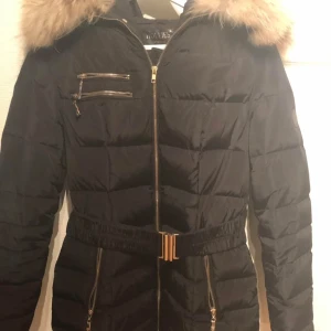 Svart Hollies jacka med pälsdetaljer  - Snygg svart Holliesjacka med dragkedja och pälsdetaljer på huvan. Jackan har ett bälte i midjan för en figursydd passform och flera praktiska fickor med dragkedjor.