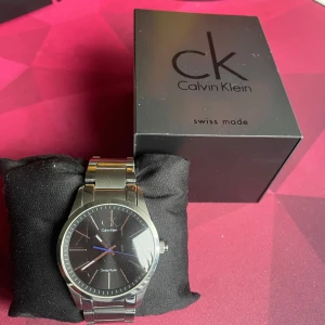 Silverfärgad klocka från Calvin Klein. Batteriet är nyss bytt - Snygg och stilren klocka från Calvin Klein i silverfärgat rostfritt stål. Den har en svart urtavla med minimalistisk design och blå sekundvisare. Klockan är Swiss Made och vattentålig upp till 10 bar. Perfekt för den som gillar en klassisk och elegant stil.    Små repor i glaset som endast syns om man kollar nära. Ligger några fler band delar i lådan. Batteriet är nyss bytt. 
