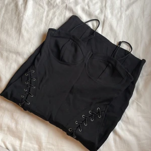 Svart klänning med snörning - Snygg svart klänning med smala axelband och snörningsdetaljer längs sidorna. Perfekt för en stilren och modern look.