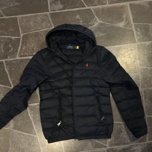 Jacka från Ralph lauren - Snygg marinblå dunjacka från Polo Ralph Lauren med huva och dragkedja. Jackan har en broderad röd logotyp på bröstet och praktiska fickor med dragkedjor. Perfekt för kyliga dagar.