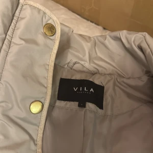 Beige quiltad jacka från Vila - Snygg beige quiltad jacka från Vila med dragkedja och knappar. Jackan har långa ärmar och praktiska fickor med dragkedja. Perfekt för kyligare dagar.