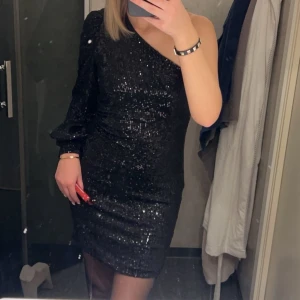 Svart glittrig klänning - Jättesnygg svart glittrig klänning med oneshoulder-design och lång ärm. Perfekt nu inför nyår 😇. Använd 1 gång så i väldigt bra skick! Pris kan diskuteras