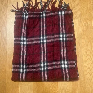 Röd Burberry halsduk  - Säljer en klassisk rutig halsduk från Burberrys i en snygg vinröd nyans med inslag av svart och vitt. Tillverkad i 100% lammull, perfekt för att hålla dig varm under kyliga dagar. Halsduken har fransar i ändarna som ger en extra touch av stil.