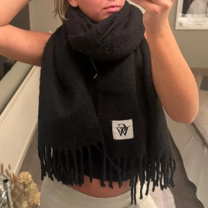 Svart halsduk med fransar - Snygg och mysig svart halsduk med fransar och en vit logotyp. Perfekt för att hålla värmen under kyliga dagar.