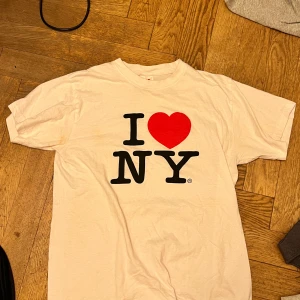 Vit 'I ❤️ NY' t-shirt - Klassisk vit t-shirt med det ikoniska 'I ❤️ NY'-trycket i svart och rött. Perfekt för alla som älskar New York! T-shirten är kortärmad och gjord i bomull, vilket gör den bekväm att bära.