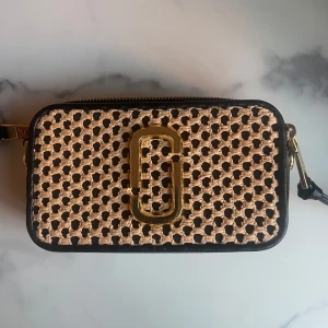 Svart och beige väska från Marc Jacobs - Snygg och unik väska från Marc Jacobs i svart och beige med ett flätat mönster. Väskan har en guldfärgad logotypdetalj på framsidan och en praktisk ficka på baksidan. Perfekt för att ge din outfit en stilfull touch.