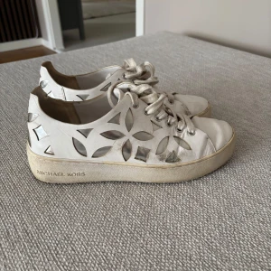 Vita sneakers från Michael Kors - Snygga vita sneakers från Michael Kors med snyggt utskuret  mönster i silver på sidorna.   Storlek 37