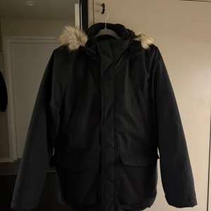 Svart parkajacka med pälsdetaljer - Snygg svart parkajacka med huva och pälsdetaljer. Jackan har långa ärmar och praktiska fickor framtill. Perfekt för kyliga dagar, väldigt varm och skön för vintern! 