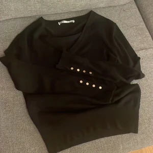 Svart tröja från Zara - Snygg svart tröja från Zara med v-ringning och dekorativa knappar på ärmarna. Perfekt för en stilren look.