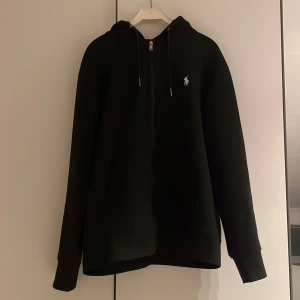 Svart hoodie från Polo Ralph Lauren - Säljer en stilren svart hoodie från Polo Ralph Lauren med dragkedja och huva. Den har en liten broderad logga på bröstet och långa ärmar. Perfekt för en avslappnad look.