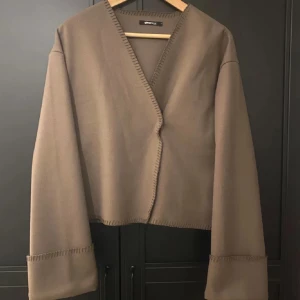 Brun kappa från ginatricot - Snygg brun kappa från ginatricot med v-ringad design och långa ärmar. Jackan har en stilren look med dekorativa sömmar längs kanterna. Perfekt för en trendig och minimalistisk stil.