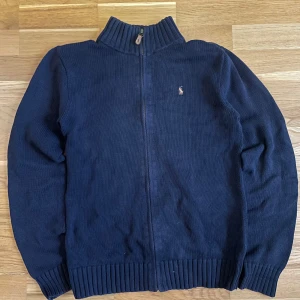 Mörkblå stickad tröja från Polo Ralph Lauren - Säljer en snygg mörkblå stickad tröja från Polo Ralph Lauren med dragkedja och broderad logga på bröstet. Tröjan har en hög krage och ribbade muddar vid ärmslut och nederkant. Perfekt för kyliga dagar! Storleken är XL 18-20 i barnstorlek 