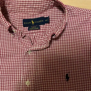 Rutig skjorta från Ralph Lauren - Snygg röd och vit rutig skjorta från Ralph Lauren i slim fit. Skjortan har långa ärmar och knappar framtill. Den är broderad med den klassiska logotypen på bröstet. Perfekt för en stilren look!