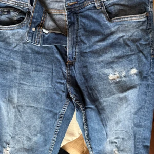 Splash DNM indigo Goods  - Säljer ett par blå jeans med coola slitningar. De har en klassisk femficksdesign och knappgylf. Perfekta för en avslappnad stil.