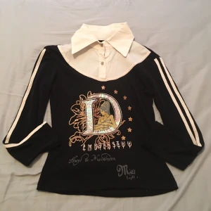 Svart och vit tröja med tryck - Snygg svart tröja med vita detaljer och ett stort tryck på framsidan. Tröjan har en unik design med en vit krage och knappar, samt vita ränder längs ärmarna. Perfekt för en cool och avslappnad stil.