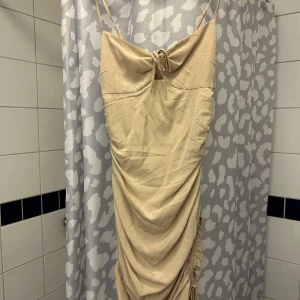 Beige klänning med rynkdetaljer - Elegant beige klänning med smala axelband och rynkdetaljer både fram och bak. Klänningen har en knytning framtill och är i en mjuk, lätt material som ger en smickrande passform.