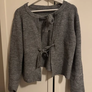 Grå stickad kofta med rosetter - Mysig grå stickad kofta med knytband framtill. Perfekt för kyliga dagar med sin ribbade struktur och långa ärmar. Aldrig använd!