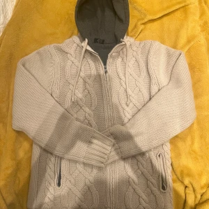 Sjukt snygg stickad cardigan - Säljer en mysig beige stickad hoodie med dragkedja och kabelstickat mönster. Tröjan har en grå huva och ribbade muddar vid ärmslut och nederkant. Perfekt för kyliga dagar!