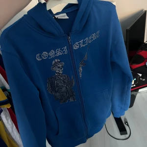 Blå hoodie med tryck - Snygg blå hoodie med dragkedja och ett coolt tryck av en figur på framsidan. Perfekt för en avslappnad stil med en touch av attityd. Huvan ger extra komfort och stil.