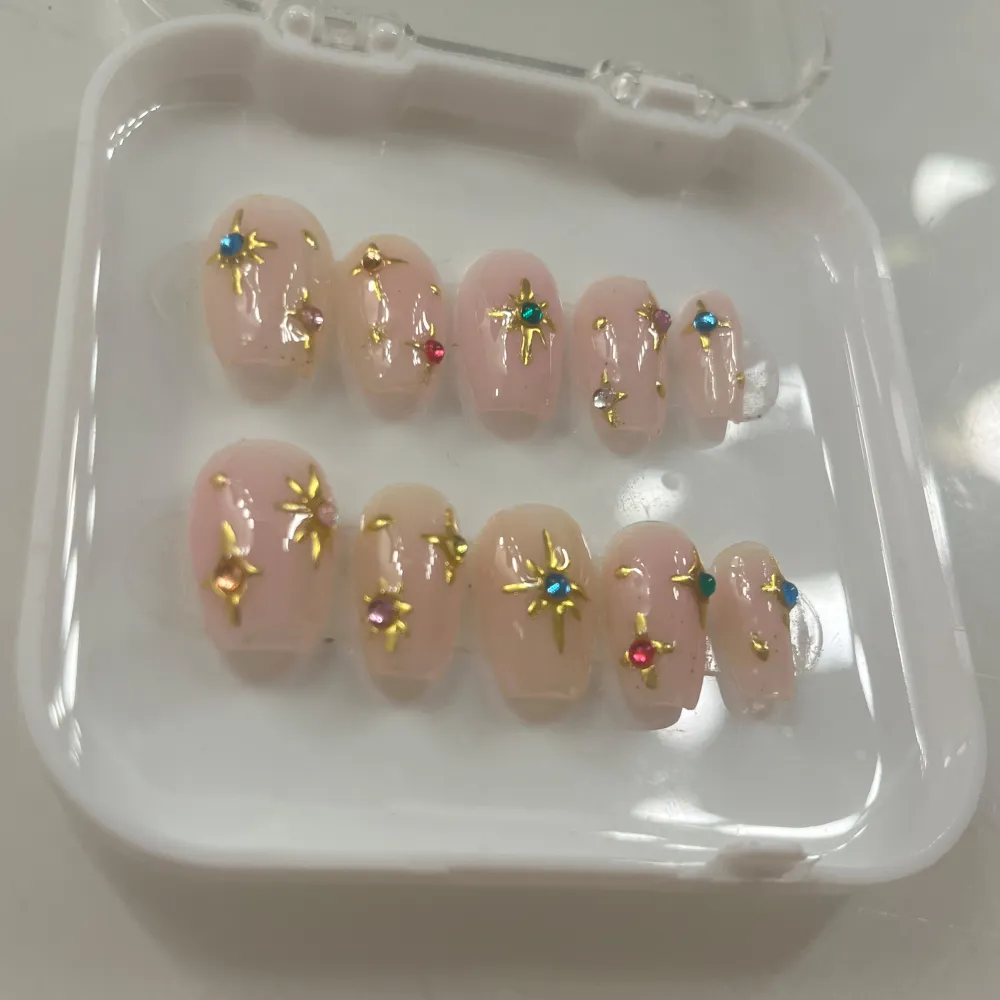 Snygga rosa lösnaglar dekorerade med guldstjärnor och färgglada stenar i blått, rött och grönt. Perfekt för en glamorös look.. Beauty.
