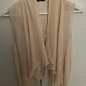 Beige väst med spetsdetaljer - Elegant beige väst med vackra spetsdetaljer på ryggen. Västen har en flödande design och är perfekt för att lägga till en romantisk touch till din outfit.