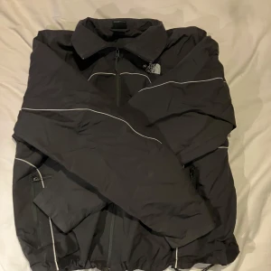 Svart vindjacka från The North Face - Säljer en svart vindjacka från The North Face med dragkedja och vita detaljer. Jackan har långa ärmar och en klassisk krage. Perfekt för blåsiga dagar.