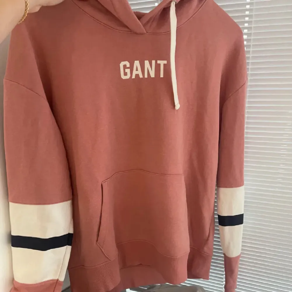 Säljer en snygg rosa hoodie från GANT med vit logga på bröstet. Tröjan har långa ärmar med vita och svarta ränder vid ärmsluten samt en praktisk magficka. Perfekt för en avslappnad stil!. Neuletakit & Villapaidat.