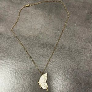 Guldhalsband med fjärilshänge - Elegant guldhalsband med ett vackert fjärilshänge i vitt. Perfekt för att lägga till en subtil touch av naturinspirerad skönhet till din stil. Kedjan är tunn och smidig, vilket gör den bekväm att bära hela dagen.