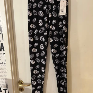 Svarta leggings med dödskallemönster - Coola svarta leggings med ett iögonfallande vitt dödskallemönster. Perfekta för en edgy look och passar bra till både vardag och speciella tillfällen. Tillverkade i ett stretchigt material för en bekväm passform.