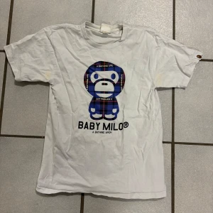 Vit Baby Milo t-shirt från A Bathing Ape - Säljer en vit t-shirt från A Bathing Ape med ett stort Baby Milo-tryck i blått och svart på framsidan. T-shirten är kortärmad och har en rund halsringning. Perfekt för dig som gillar streetwear och unika tryck! Köpt i London 