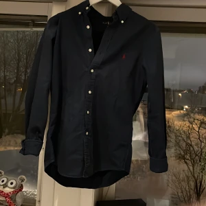 Mörkblå skjorta från Ralph Lauren - Snygg mörkblå skjorta från Ralph Lauren i slim fit. Skjortan har långa ärmar och knappar framtill. Perfekt för en stilren look. Skjorta är knappast använd.