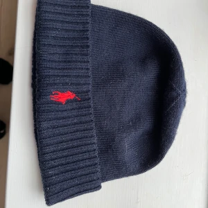 Mörkblå stickad mössa från Ralph Lauren - Snygg mörkblå stickad mössa från Ralph Lauren med röd broderad logga framtill. Perfekt för kyliga dagar och ger en stilren look. Passar både till vardags och mer uppklädda tillfällen.