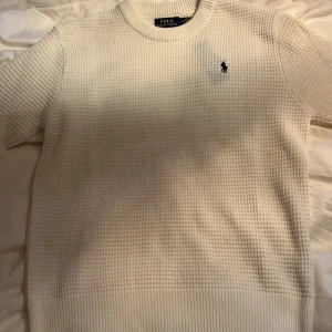 Vit tröja från Polo Ralph Lauren - Säljer en snygg och skön vit Ralph Lauren knitted med det klassiska logotypbroderiet på bröstet. Tröjan har en ribbad rund hals och är tillverkad i ett mjukt material med textur. Perfekt för en avslappnad och elegant look.