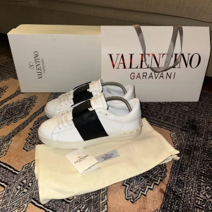 Valentino Garavani sneakers i vitt och svart - Snygga Valentino Garavani sneakers i vitt med en bred svart rem över ovansidan. Skorna har en stilren design med snörning och är perfekta för en trendig look.