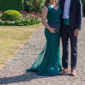 Grön långklänning med spetsdetaljer - Mörkgrön långklänning med v-ringning fram samt bak och vackra spetsdetaljer. Klänningen har en smickrande passform och ett bälte i midjan. Fina knappar i ryggen, inte upplagd, använd 1 gång sedan har den tvättats. Väldigt fint skick och har ett fantastisk släp! Inköp på Dressroom Linne för 2900kr - orginal klädpåse medföljer