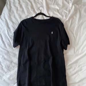 Svart t-shirt från Ralph Lauren - Säljer en klassisk svart t-shirt från Ralph Lauren med det ikoniska broderade logotypen på bröstet. T-shirten är kortärmad och tillverkad i mjuk bomull för en bekväm passform. T-shirten är aldrig använd.
