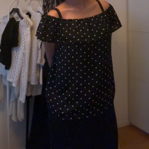 Off shoulder blus - Snygg svart blus med vita prickar och offshoulder-design. Perfekt för en stilren look med en touch av lekfullhet. Klänningen har en luftig passform och är ärmlös med volangdetaljer vid axlarna.