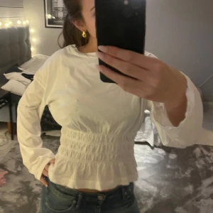 Vit tröja  med volangdetalj - Säljer en söt vit tröja med volangdetalj i midjan. Blusen har långa ärmar och en smockad midja som ger en fin passform. Perfekt för en stilren look.