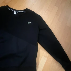 Svart sweatshirt från Lacoste - Säljer en stilren svart sweatshirt från Lacoste med det klassiska krokodilmärket på bröstet. Tröjan har långa ärmar och en rund halsringning med vit randdetalj. Perfekt för en avslappnad look.